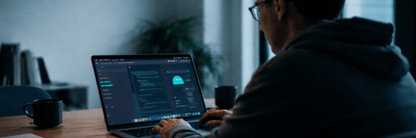 Programmierer arbeitet konzentriert an einem Laptop mit einem KI-Assistenten in moderner, ruhiger Arbeitsumgebung; warme Lichtstimmung, unscharfer Hintergrund mit Pflanze und Tasse auf dem Tisch